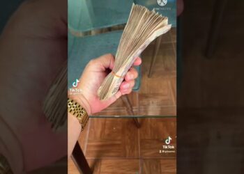 Mano con reloj dorado sosteniendo un fajo de billetes sobre una mesa de vidrio, ¿Cuánto cobra BXS por sus presentaciones en eventos privados?