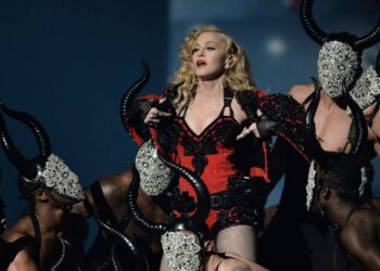 Artista en escena con traje rojo y negro rodeada de bailarines con máscaras ornamentadas y cuernos, ¿Cuánto cobra Tyga por concierto? Descubre su caché actual