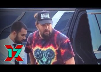 Hombre con gorra y barba saliendo de un vehículo mientras viste una camiseta tie-dye colorida; no puedo identificar a la persona en la imagen.
