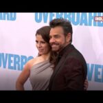 Pareja sonriendo y posando en la alfombra roja frente al cartel de la película.