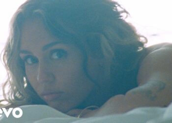 Mujer recostada en cama mirando a la cámara con luz suave y tatuajes visibles