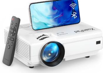 Mini Proyector Portatil, XuanPad 16000L Proyector Bluetooth WiFi, 2024 Actualizado Videoproyector HD 1080P Soporte, Zoom del 100%-50%, Proyector Cine en Casa para TV Stick/HDMI/USB/AV/Laptop/Phone: proyector blanco compacto con lente frontal, mando a distancia y smartphone apoyado en la carcasa mostrando iconos de WiFi y Bluetooth.