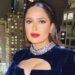 Actriz o celebridad en balcón nocturno con vestido azul de terciopelo, collar de brillantes y labios rojos, vista urbana iluminada