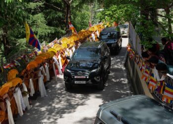 Monjes con sombreros amarillos y público alineado junto a una caravana de vehículos entre banderas tibetanas en Dharamsala, Dónde vive el Dalai Lama: descubre su hogar en Dharamsala.