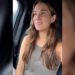 Mujer joven sonriente sentada en el asiento delantero de un coche, mirando hacia la ventana con luz natural.