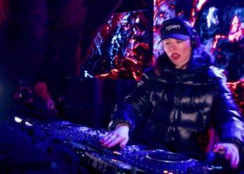 DJ femenina con gorra y abrigo negro mezclando en consola iluminada; ¿Cuánto cobra Nina Kraviz por sus shows? Descubre su tarifa real
