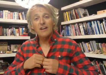 Hombre con camisa de cuadros rojos y verdes frente a estanterías llenas de libros en una biblioteca doméstica, ambiente acogedor y personal.