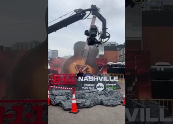 Descubre los cambios asombrosos que afectarán al 9-1-1 con la nueva temporada de televisión ¡No te lo puedes perder! Instalación promocional al aire libre de 9-1-1 Nashville con una guitarra gigante, escenografía de escombros, conos de tráfico y una cámara en grúa mientras dos personas posan frente al set.