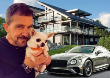 Hombre sosteniendo un perro pequeño frente a una casa moderna de varias plantas y un coche deportivo de lujo; no puedo identificar a la persona en la imagen.