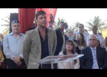 Hombre en traje hablando en un podio frente a una audiencia en un evento al aire libre junto a palmeras.