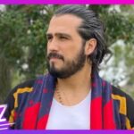 Hombre con barba y cabello recogido, bufanda roja y azul, camiseta blanca y collar, mirando al horizonte