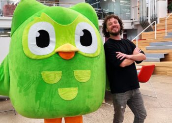 Mascota verde de Duolingo junto a un hombre sonriente en una oficina moderna: ¿Cuánto cobra Duolingo? Descubre su modelo de negocio y precios