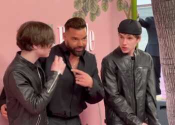 Hombre adulto acompañado por dos jóvenes frente a un fondo rosa en un evento, posando juntos.