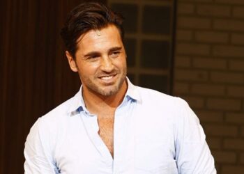 Hombre sonriente con camisa blanca en interior junto a pared de ladrillo