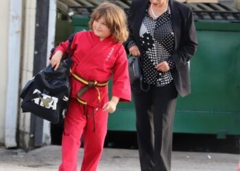 Niño con kimono rojo y cinturón marrón cargando una bolsa deportiva junto a una mujer adulta en la calle