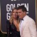 Pareja posa en la alfombra roja de los Golden Globes; ella con vestido negro brillante y joyería, él con esmoquin blanco