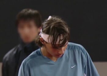 Tenista concentrado en la cancha con cinta blanca en la frente y camiseta deportiva azul, gesto pensativo durante el partido.