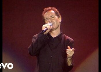 Cantante masculino en escenario con micrófono y chaqueta oscura, interpretando ante iluminación cálida.