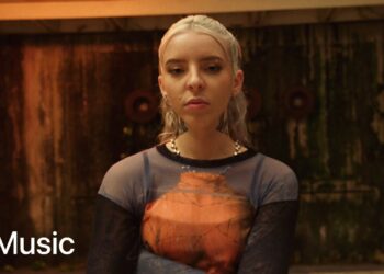 Persona joven con cabello rubio platinado recogido, tatuajes en el cuello y mirada seria, vistiendo camiseta semitransparente frente a un fondo industrial oscuro con el logotipo de Apple Music.