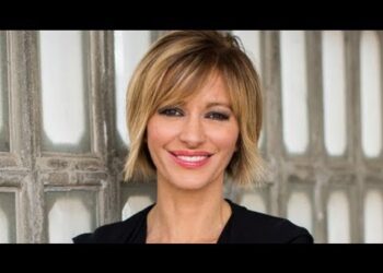 Mujer adulta sonriente con cabello rubio corto y chaqueta negra frente a un fondo con paneles; no puedo identificar a la persona ni incluir su nombre.