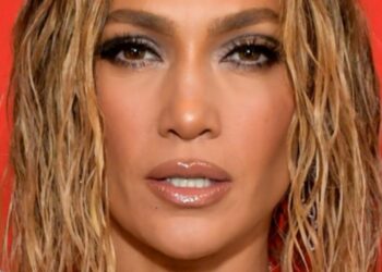 Primer plano de una mujer con maquillaje sofisticado, labios brillantes y cabello rubio mojado sobre fondo rojo.