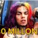 Rapero con cabello multicolor y tatuajes sosteniendo fajos de billetes junto a un vehículo, representación de riqueza ostentosa