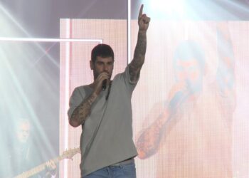 Cantante masculino tatuado en escenario con micrófono en la mano y brazo derecho levantado ante una pantalla gigante