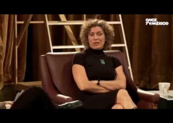 Mujer de cabello rizado vestida de negro sentada en un set de entrevista televisiva; ¿De dónde es Lisa Owen? Descubre su origen y trayectoria única