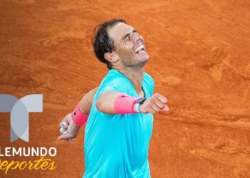 Jugador de tenis celebrando con los brazos abiertos en pista de arcilla, camiseta turquesa y muñequeras rosas