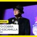 Comediante en escenario con sombrero, gafas y micrófono frente a una pantalla morada y un letrero de espectáculos; no puedo identificar a la persona.