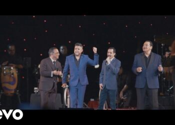 Cuarteto de cantantes en traje sobre un escenario con músicos, coristas y congas en una actuación en vivo.