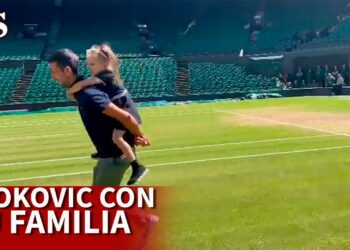 Hombre corriendo por la pista de césped con una niña a la espalda en un estadio de tenis, mostrando un instante íntimo de vida familiar.