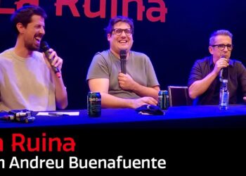 Tres presentadores sonrientes sentados en una mesa con micrófonos, risas y ambiente de comedia en un escenario iluminado