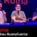 Tres presentadores sonrientes sentados en una mesa con micrófonos, risas y ambiente de comedia en un escenario iluminado