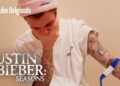 ¿De dónde es Justin Bieber? Descubre sus raíces canadienses 13 Imagen de un artista con gorro rosa, camiseta blanca y brazos tatuados, mano cubriendo el rostro; ¿De dónde es este artista? Descubre sus raíces canadienses