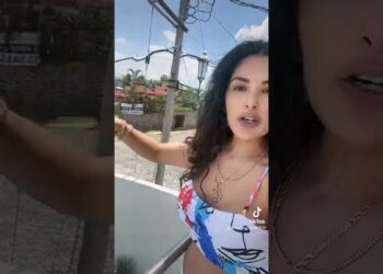 Mujer en un balcón grabando un video vertical junto a postes eléctricos, palmeras y casas al fondo; ¿Dónde trabaja Eduardo Trelles? Descubre su nueva etapa profesional.
