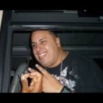 Hombre sonriente dentro de un vehículo haciendo un gesto con las manos, con camiseta oscura, cadena y reloj