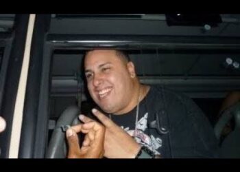 Hombre sonriente dentro de un vehículo haciendo un gesto con las manos, con camiseta oscura, cadena y reloj