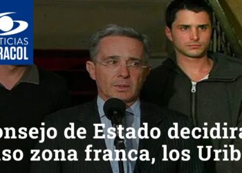 Hombre mayor hablando frente a un micrófono, flanqueado por dos hombres jóvenes y logo de Noticias Caracol en la esquina