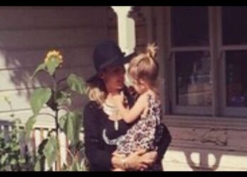 Mujer con sombrero abrazando a una niña frente a una casa, escena familiar y cálida