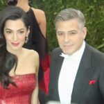 Pareja elegante en alfombra roja: hombre con esmoquin negro y pajarita blanca junto a mujer con vestido rojo brillante y cabello suelto