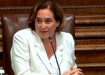 Mujer sentada en un sillón de madera con micrófono, vestida con chaqueta blanca, en interior institucional