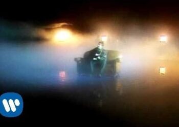 Persona sentada en un sillón rodeada de humo y luces cálidas con un logo discográfico visible