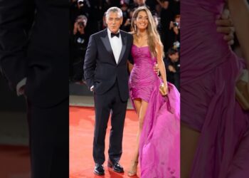 Pareja en alfombra roja: mujer sonriente con vestido fucsia corto con larga cola y abertura lateral, bolso dorado y sandalias, acompañada por hombre con esmoquin negro.