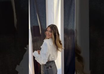 Mujer joven sonriendo hacia la cámara mientras se vuelve junto a unas cortinas, con blusa blanca y vaqueros de talle alto.