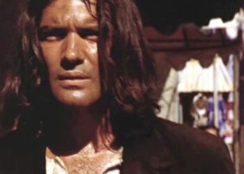 Primer plano de un actor con cabello largo y mirada intensa en un entorno urbano, iluminación cálida y vestuario de época