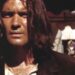 Primer plano de un actor con cabello largo y mirada intensa en un entorno urbano, iluminación cálida y vestuario de época