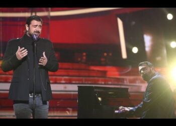 Cantante masculino interpretando una canción frente a un micrófono acompañado por un pianista al piano en un escenario iluminado; no se identifica a las personas.