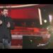 Cantante masculino interpretando una canción frente a un micrófono acompañado por un pianista al piano en un escenario iluminado; no se identifica a las personas.