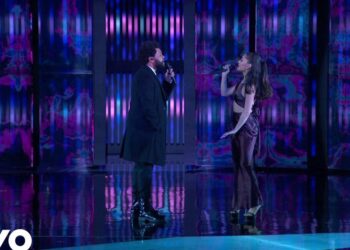 Dos cantantes en escenario con iluminación púrpura y azul durante una emotiva actuación en vivo, ambos sosteniendo micrófonos.